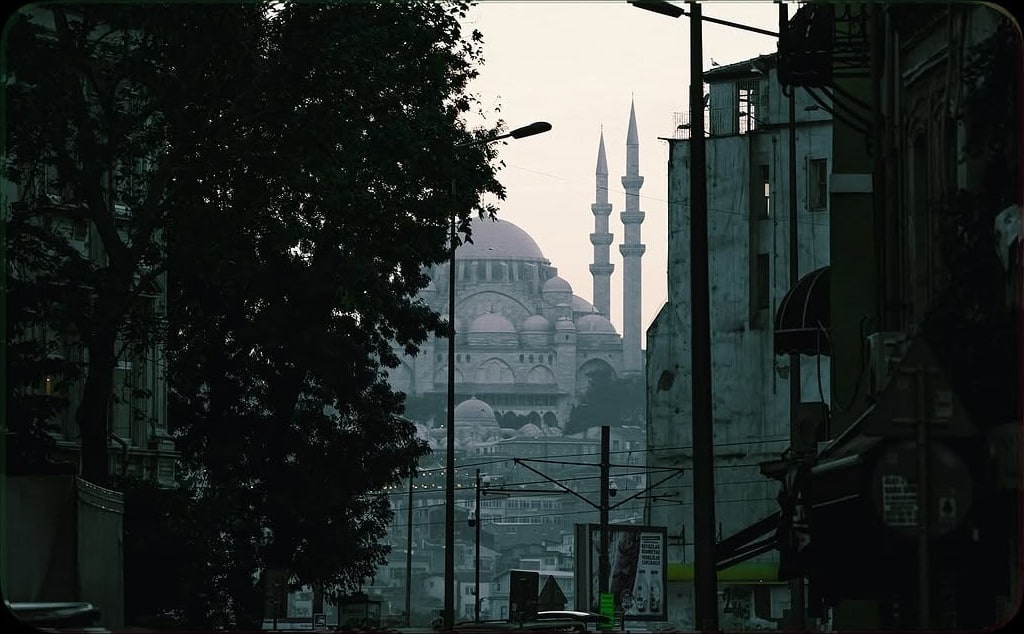 A misty mosque and its minarets glimpsed down a dark tree-lined city street at dawn.