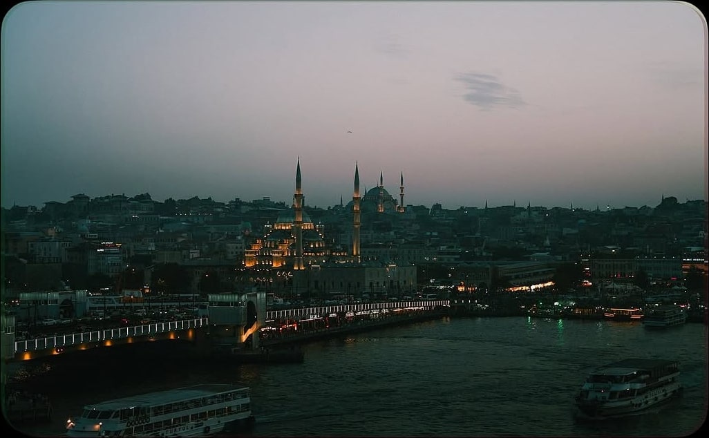 A dusky waterfront panorama with an illuminated mosque, the Galata Bridge, and ferries on dark water.