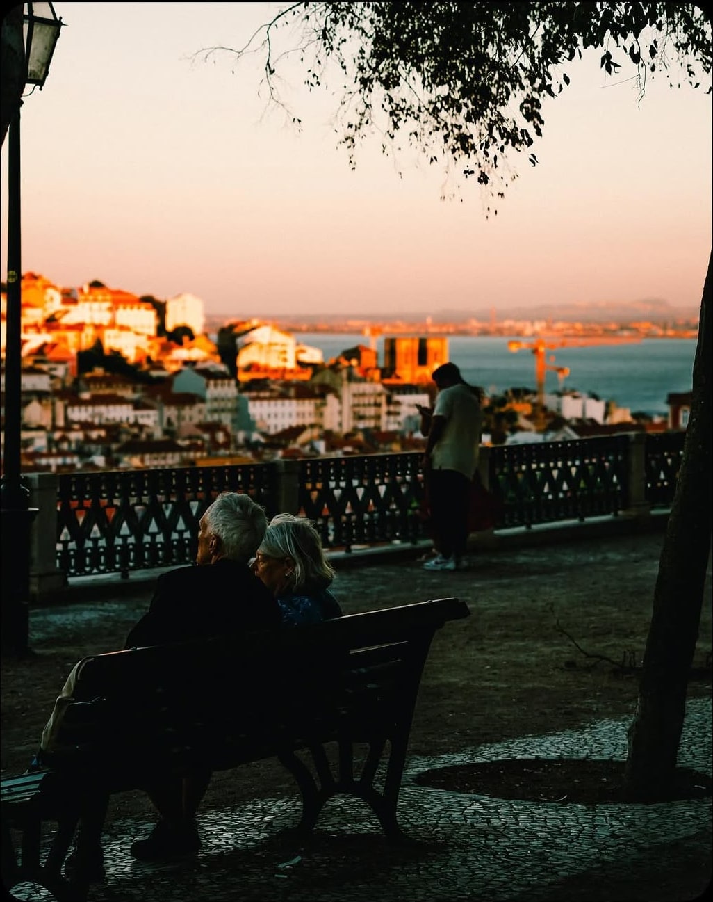 An elderly couple sits on a bench at a miradouro overlooking Lisbon rooftops at sunset.