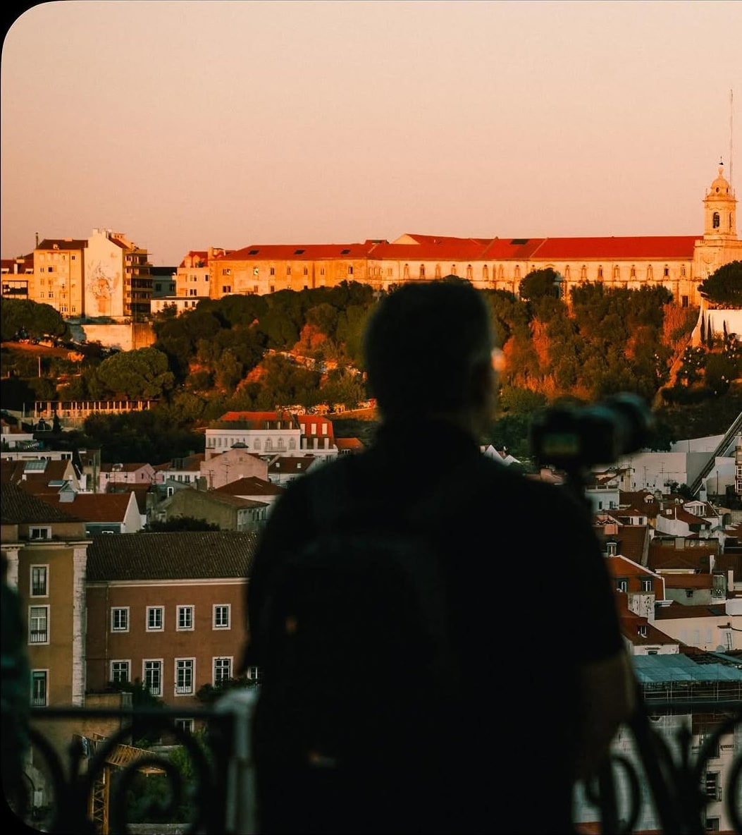 A man with a backpack and camera gazes over terracotta rooftops toward a sunlit hilltop convent.