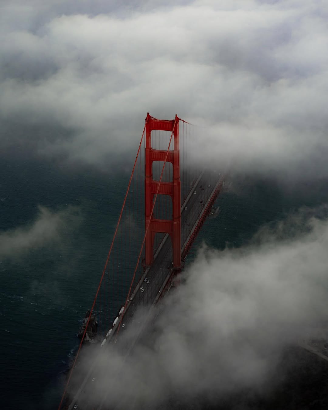 Aerial view of a Golden Gate Bridge tower piercing a bank of fog, with cars crossing the deck over dark water.