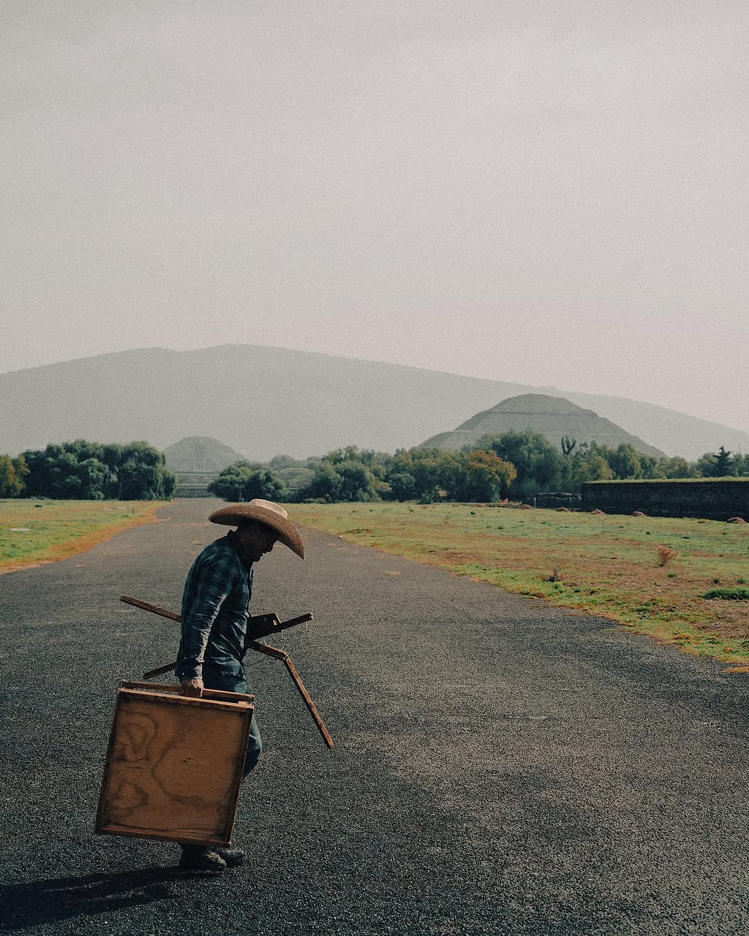 A man in a cowboy hat carries a wooden table and folded chairs across an empty road, hazy pyramids of Teotihuacan behind.