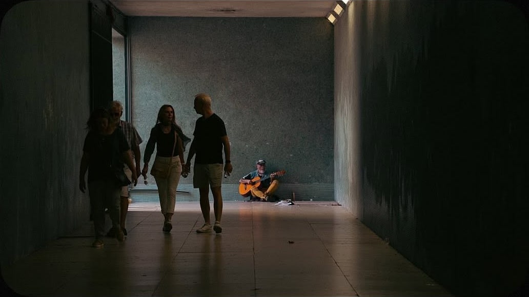 A busker plays guitar seated at the far wall of a dim tiled corridor as four pedestrians walk past.