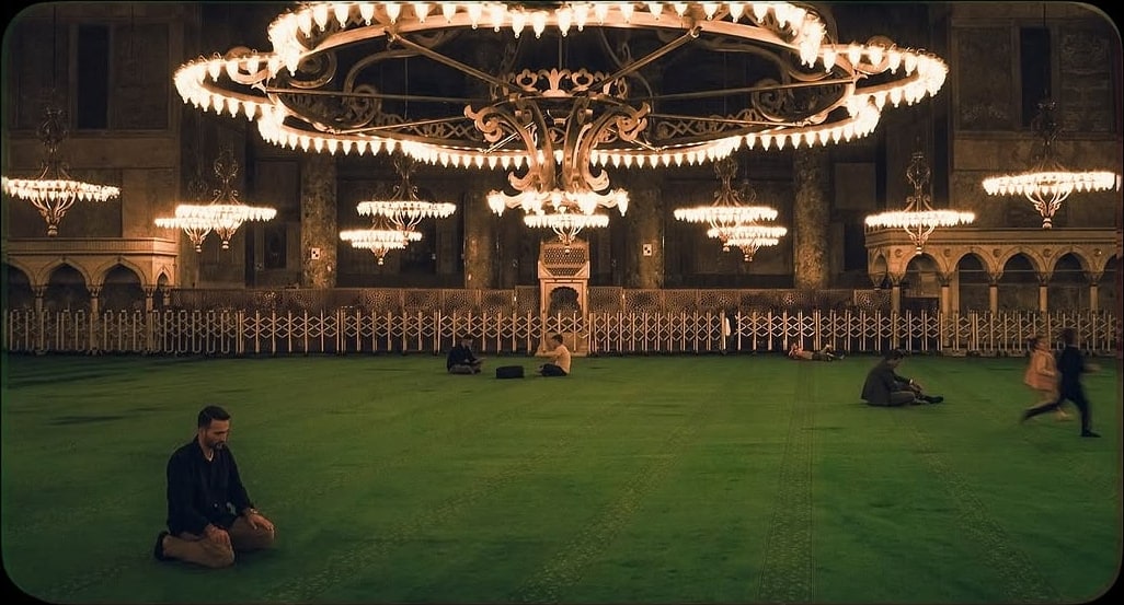 A man kneels in prayer on the green carpet of Hagia Sophia under enormous circular chandeliers.