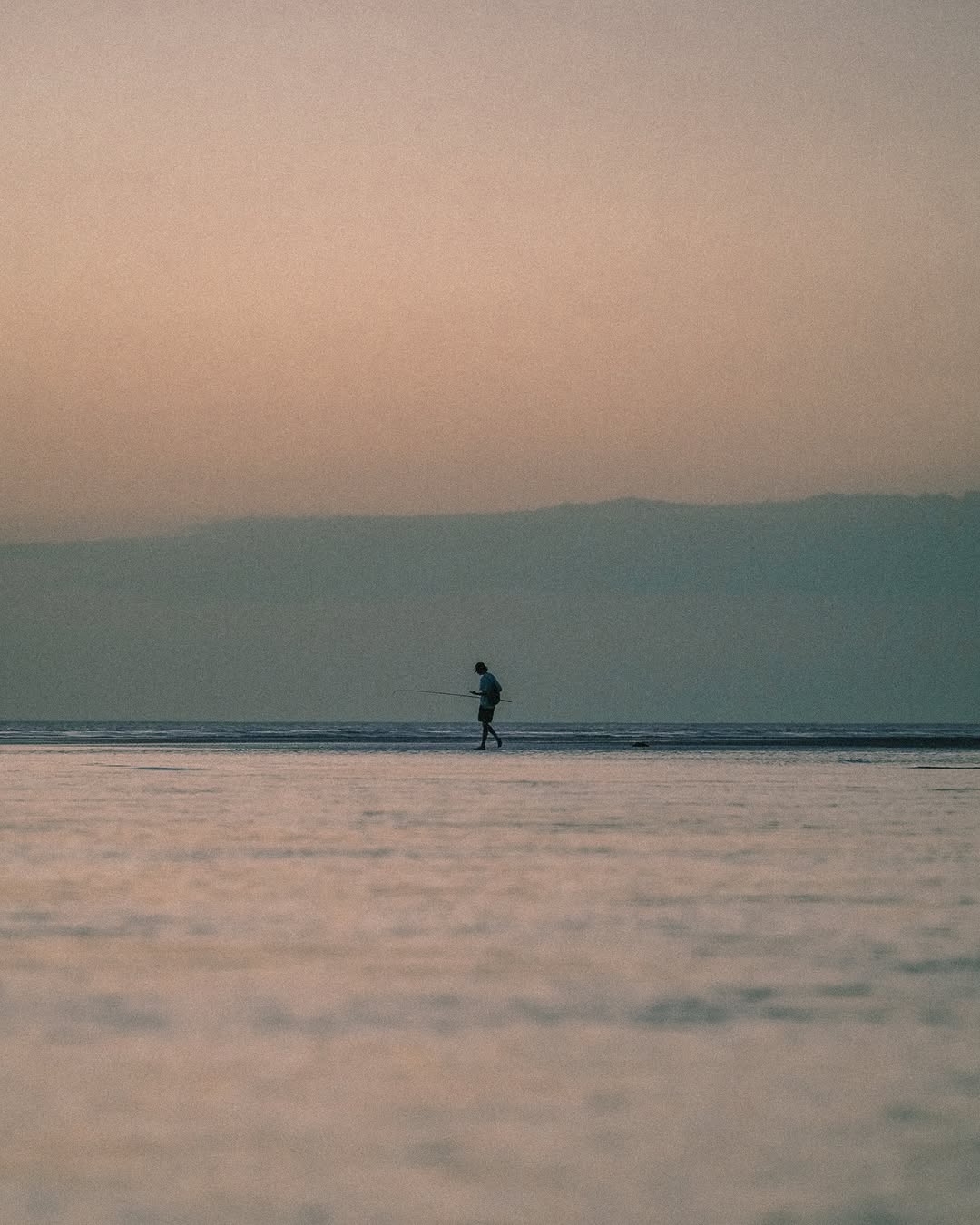 A silhouetted man carries a fishing rod across a tidal flat under a soft pink and blue pastel sky.