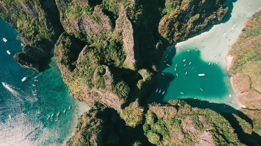 Top-down view of jungle-covered karst cliffs wrapping around a turquoise lagoon dotted with longtail boats.