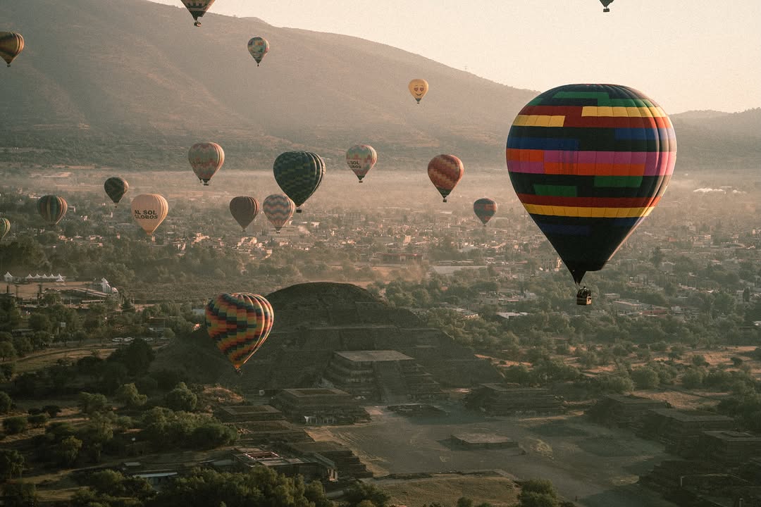 Dozens of colorful hot air balloons drift over the Teotihuacan valley and pyramid at hazy sunrise.