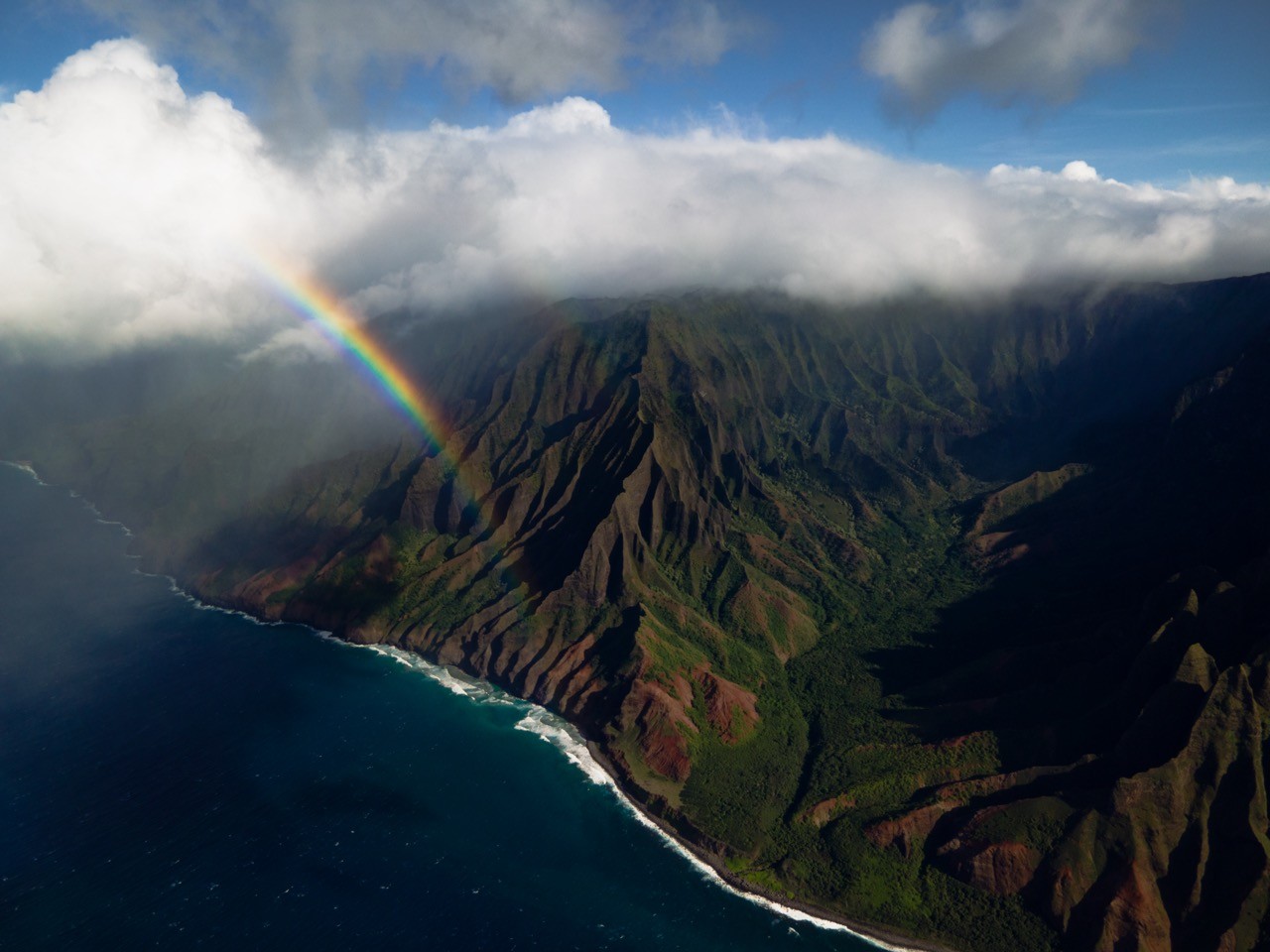 The ridges of the Nā Pali coast shot from a drone, with a rainbow arcing over the coastline.